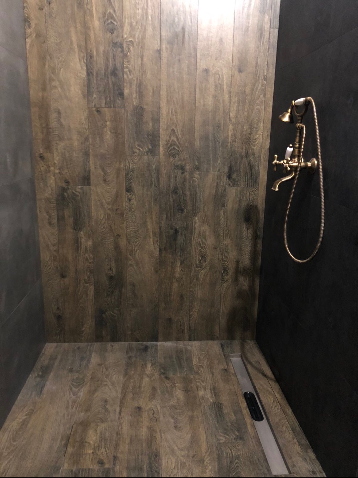 Nowoczesna kabina prysznicowa z ciemnymi płytkami imitującymi drewno, złotą armaturą i odpływem liniowym. Minimalistyczny design w łazience.