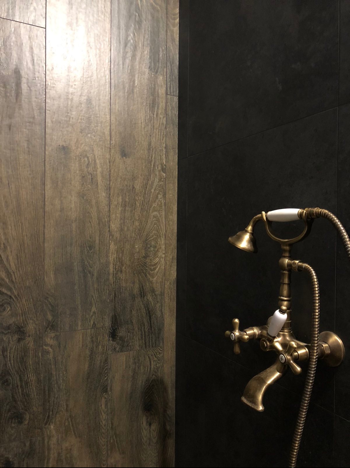 Elegancka łazienka z drewnopodobnymi płytkami i czarną ścianą, na której zamontowano stylową, mosiężną baterię z prysznicem retro. Kontrast faktur i kolorów.