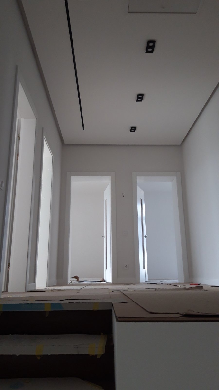 Wnętrze w trakcie wykańczania: białe ściany, sufit z oświetleniem szynowym i punktowym, otwarte drzwi. Podłoga zabezpieczona kartonem, widoczne schody w tle.