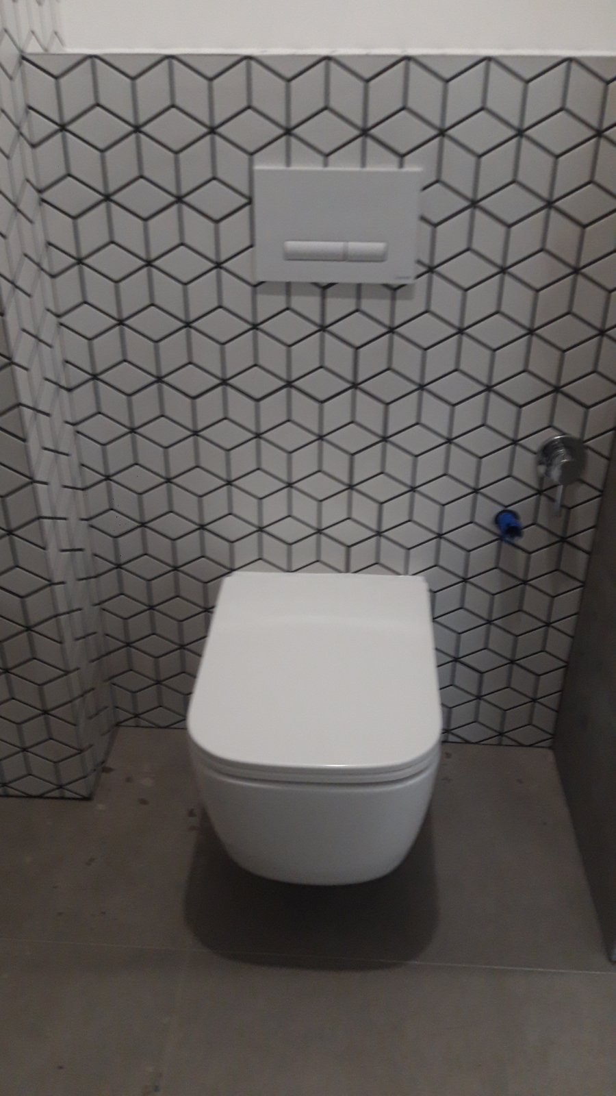 Nowoczesna toaleta wisząca na tle ściany z geometrycznych płytek w kształcie sześcianów. Widoczny przycisk spłuczki i fragment armatury.