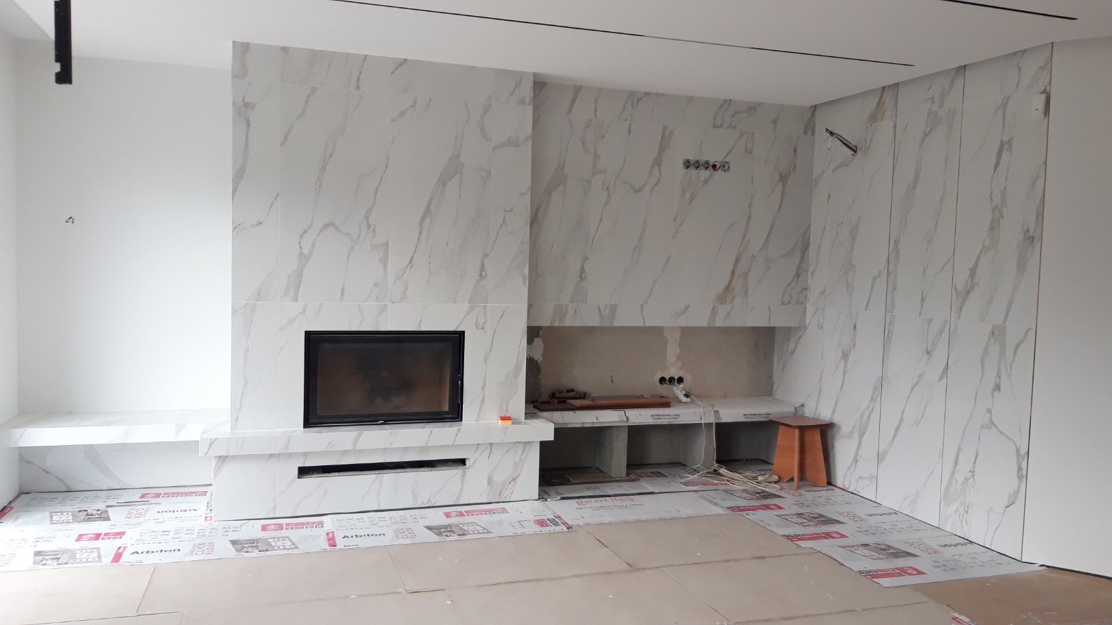 Wykończony kominek z marmurowymi płytkami w jasnym salonie; widoczne przygotowania do dalszych prac wykończeniowych.