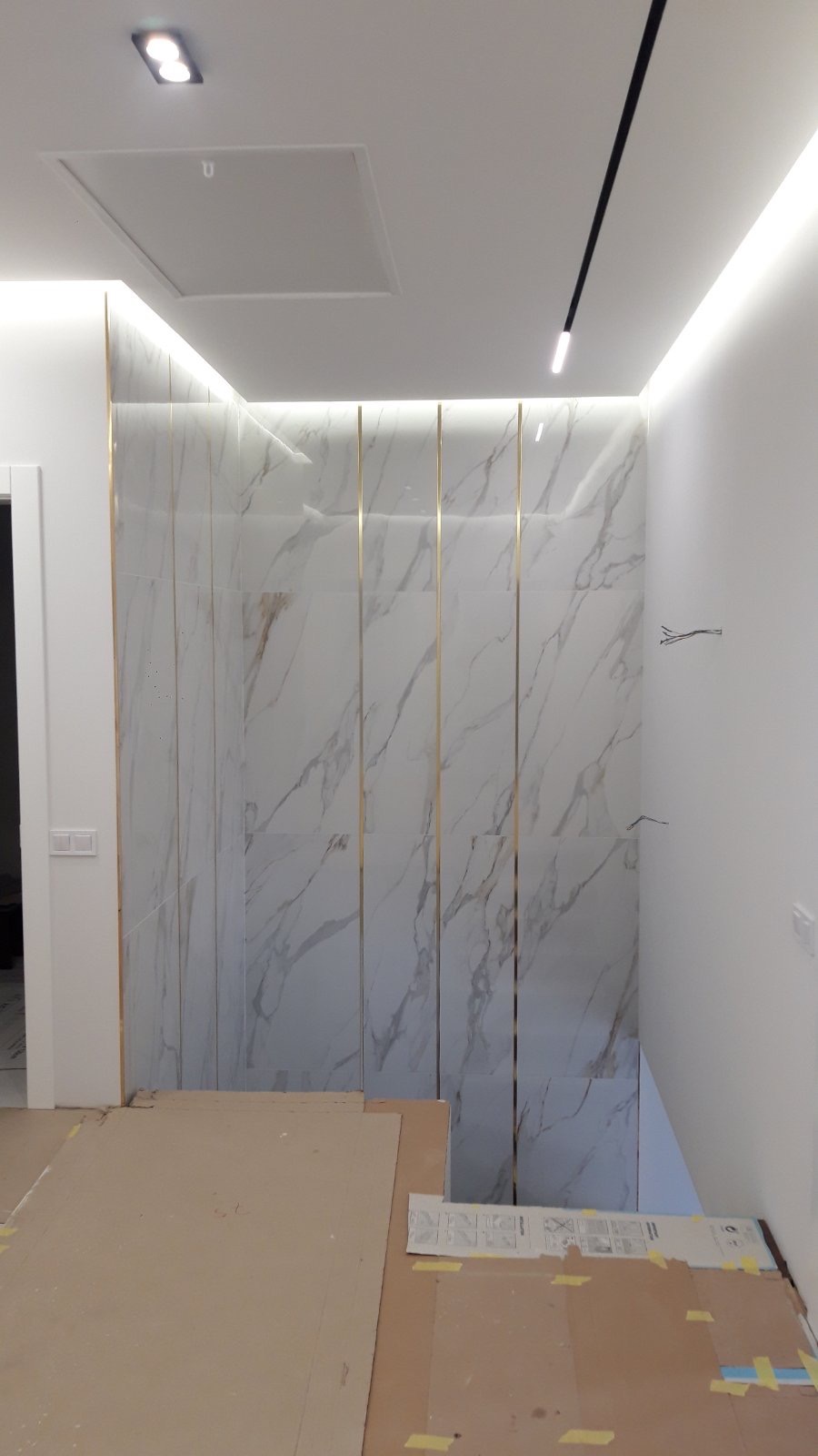 Ściana wykończona płytami imitującymi marmur, oddzielonymi złotymi listwami. Podłoga zabezpieczona kartonem. Widoczne oświetlenie sufitowe.