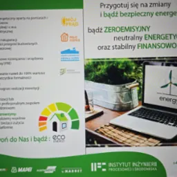 Reklama firmy oferującej audyt energetyczny i rozwiązania związane z odnawialnymi źródłami energii, z laptopem wyświetlającym logo wiatraka i hasło 'energy' w tle, obok metalowego pojemnika...