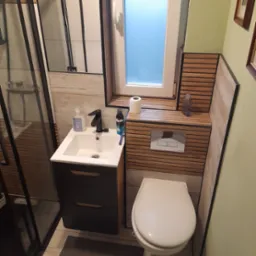 Nowoczesna łazienka po remoncie: kabina prysznicowa typu walk-in, umywalka z szafką, toaleta podwieszana. Drewniane panele dekoracyjne na ścianie i pod oknem.