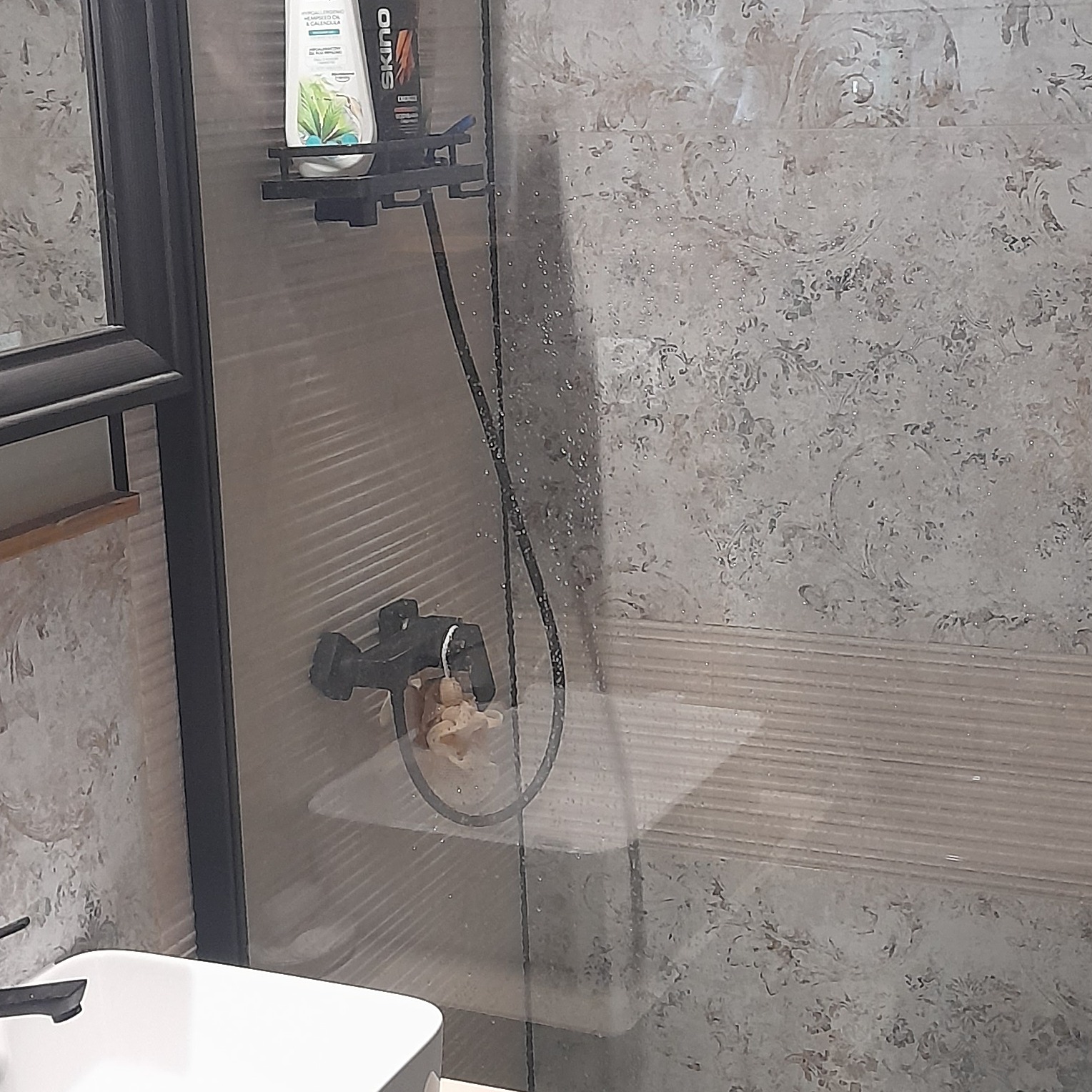 Szklana kabina prysznicowa z czarną armaturą, widoczne płytki z dekoracyjnym wzorem, półka z kosmetykami i gąbka do mycia.