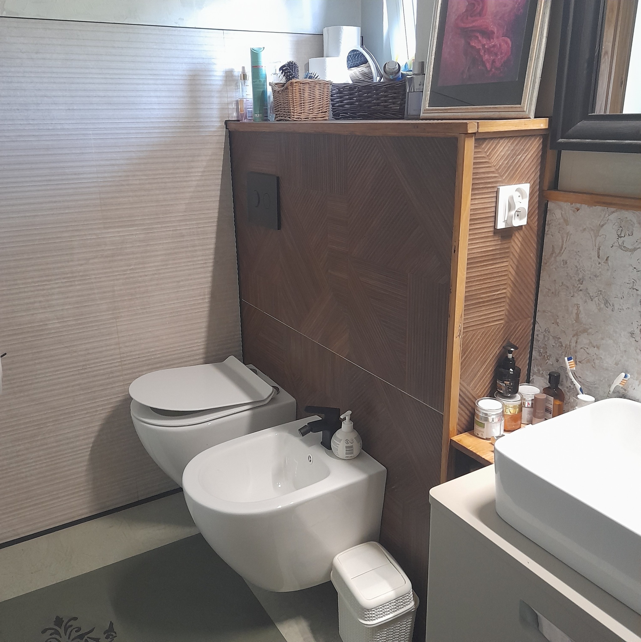 Nowoczesna łazienka z wiszącą toaletą i bidetem, wykończona płytkami w odcieniach beżu i brązu, z drewnianymi akcentami i minimalistycznym wyposażeniem.