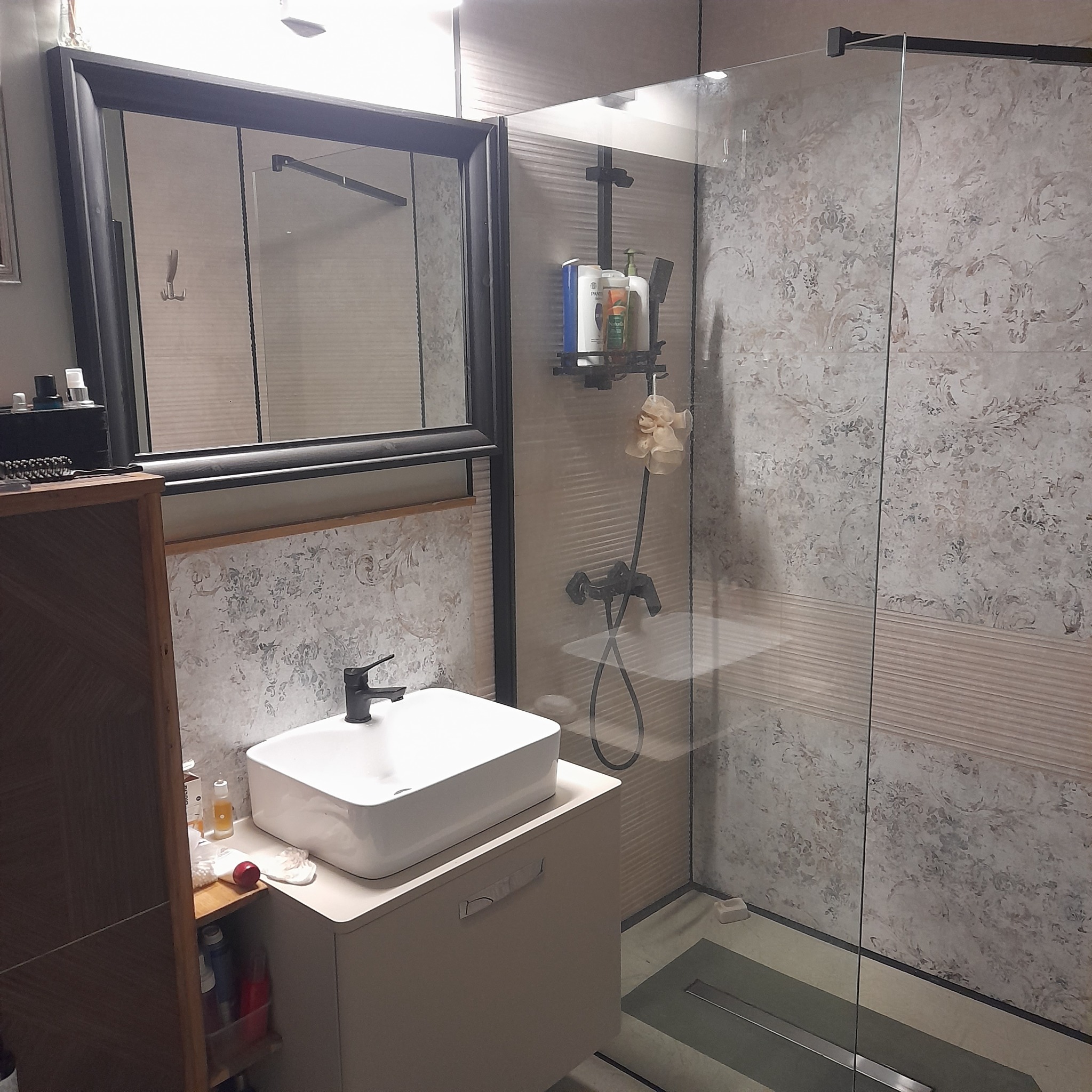 Nowoczesna łazienka z kabiną prysznicową typu walk-in, umywalką nablatową na szafce z frezowanymi uchwytami, czarną armaturą i dekoracyjnymi płytkami ściennymi w odcieniach beżu i szarości.