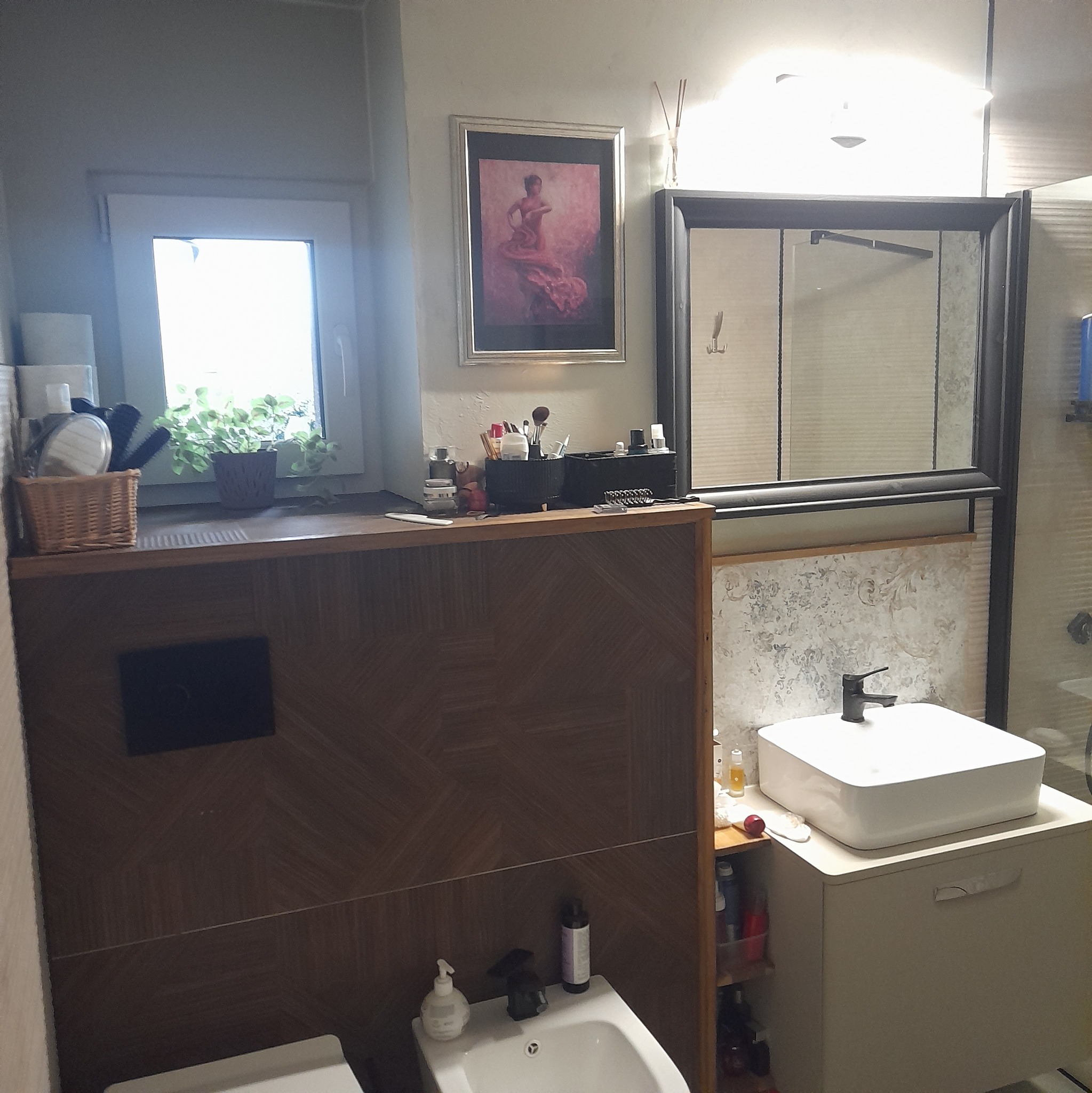 Wykończona łazienka z umywalką nablatową, bidetem, kabiną prysznicową z czarną ramą i oknem z roślinami, ozdobiona obrazem tancerki flamenco.