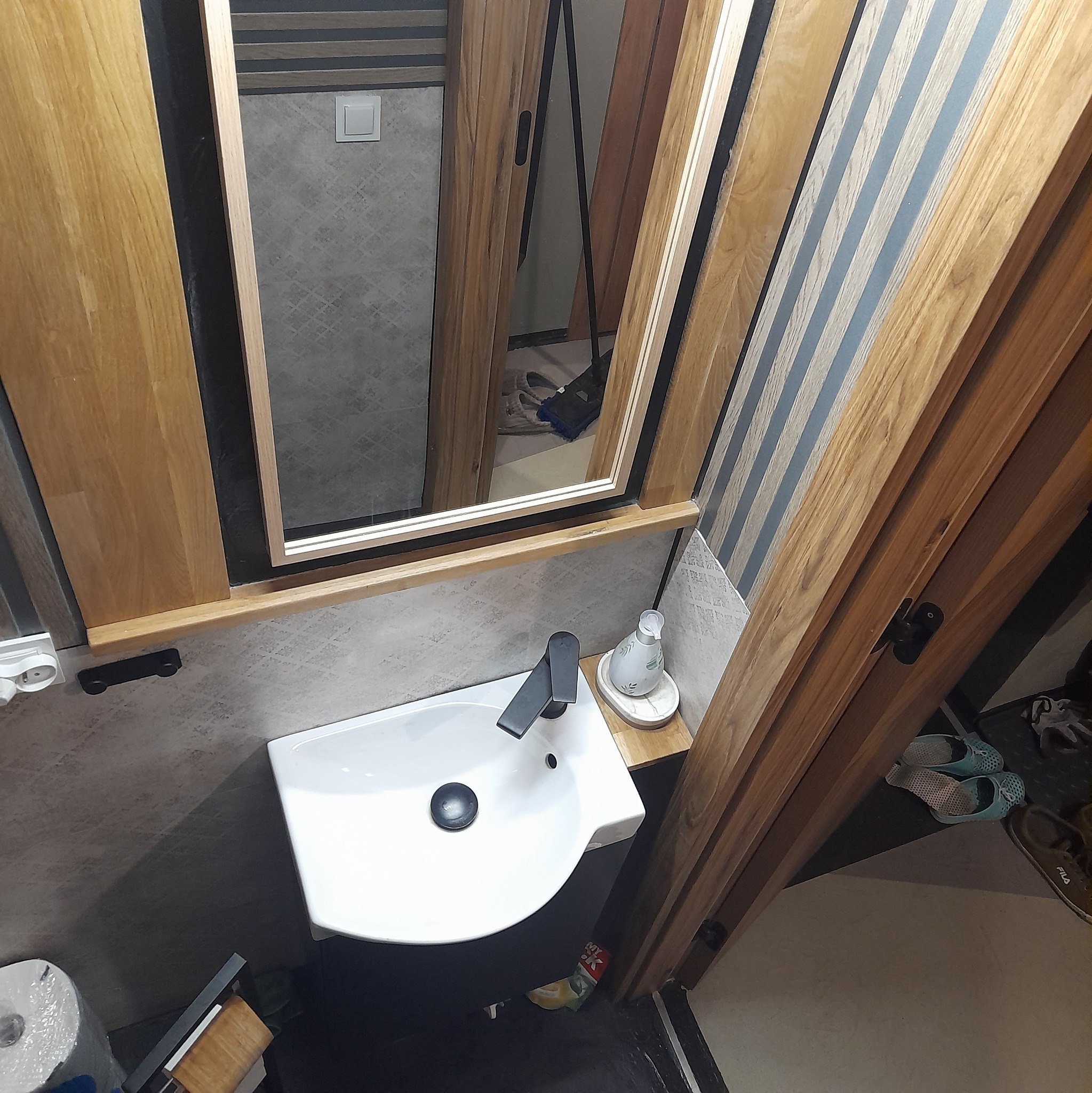 Umywalka nablatowa z czarną armaturą, biała butelka z dozownikiem na drewnianym blacie, lustro w drewnianej ramie z odbiciem fragmentu pomieszczenia, szaro-biała ściana, drewniane panele ścienne...