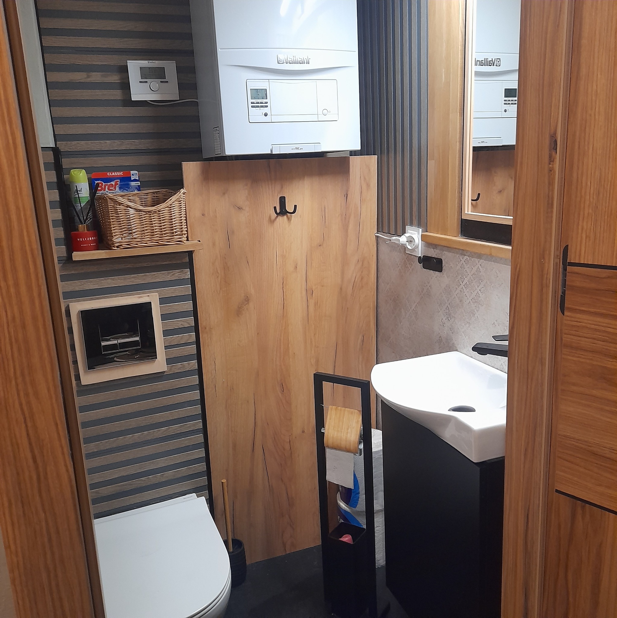 Nowoczesna łazienka po remoncie: biała umywalka z czarną szafką, wiszący piec gazowy Vaillant nad drewnianą zabudową z haczykiem, toaleta, dekoracyjne płytki ścienne i stojak na papier toaletowy.