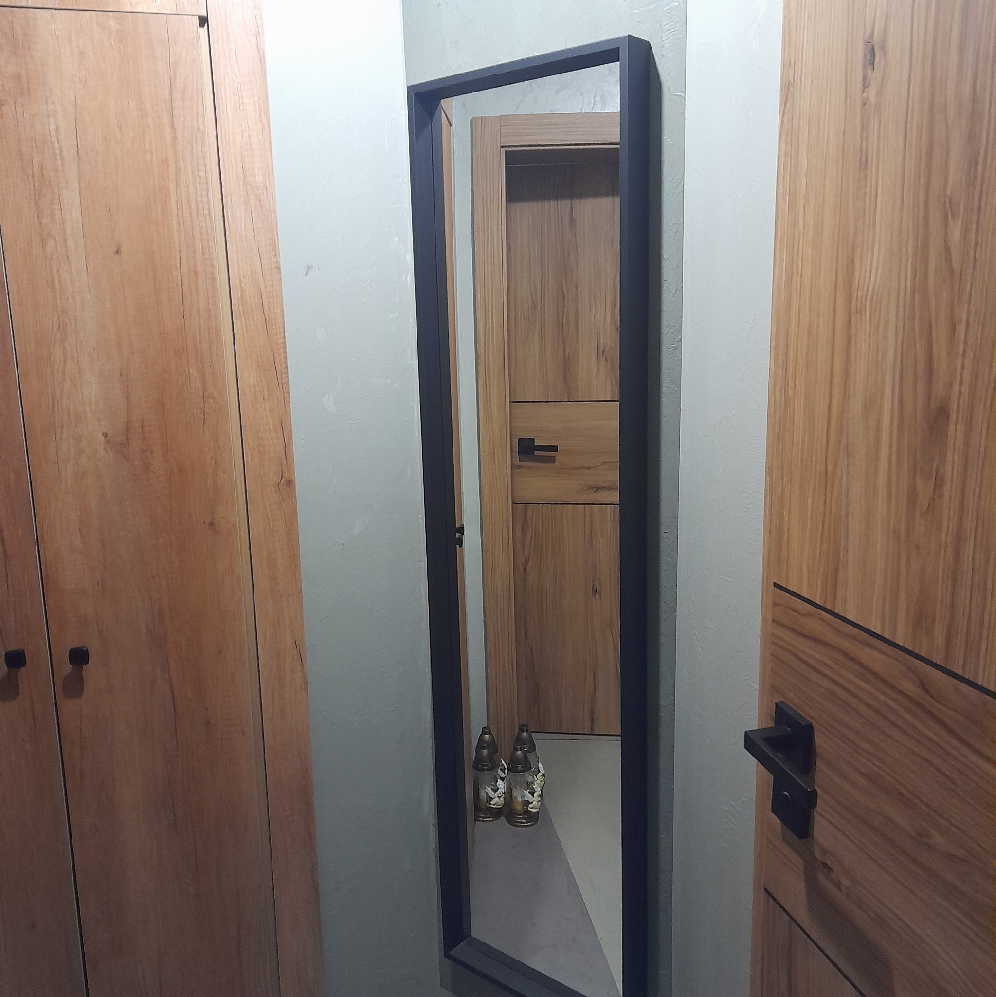 Wąskie lustro w czarnej ramie zawieszone na szarej ścianie, odbijające fragment drewnianej zabudowy i dwa znicze na podłodze.