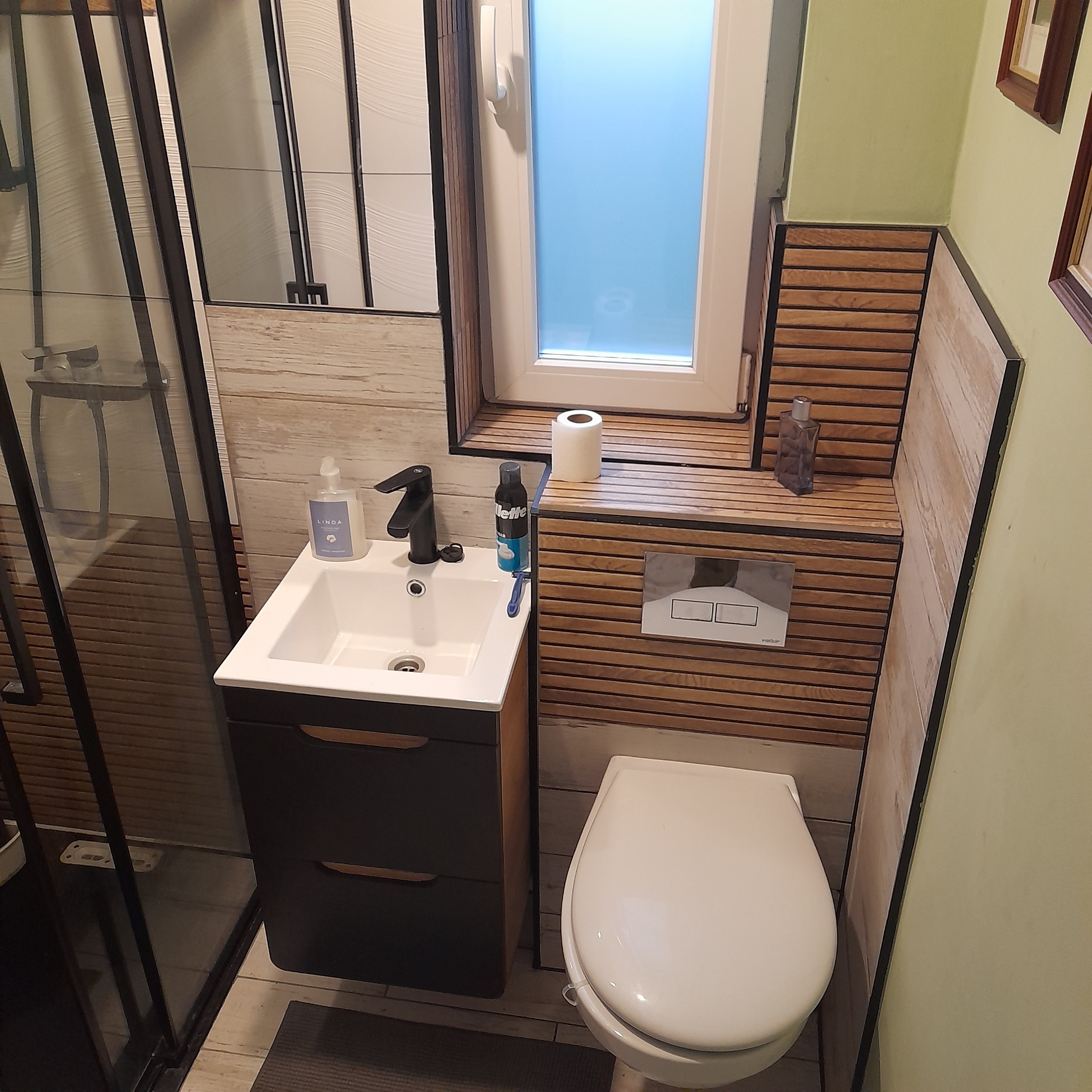 Nowoczesna łazienka po remoncie: kabina prysznicowa typu walk-in, umywalka z szafką, toaleta podwieszana. Drewniane panele dekoracyjne na ścianie i pod oknem.