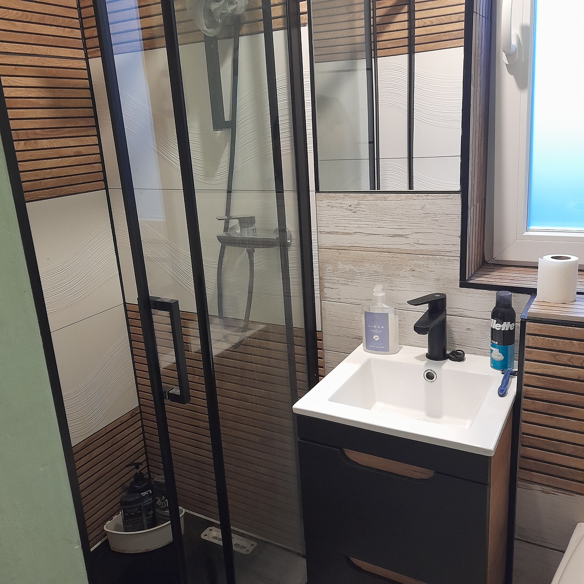 Nowoczesna łazienka z kabiną prysznicową typu walk-in, umywalką z szafką w kolorze czarnym i drewnianymi akcentami na ścianach oraz lustrze.