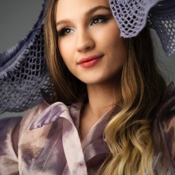 Martyna Mierzicka - MARLENS Fotografia - Portret młodej kobiety z delikatnym makijażem, ubrana w prześwitującą fioletową bluzkę i ażurową chustę na głowie, spoglądającej w górę