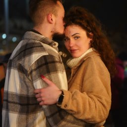 Martyna Mierzicka - MARLENS Fotografia - Mężczyzna w koszuli w kratę całuje kobietę w brązowym płaszczu w czoło; kobieta patrzy w obiektyw.