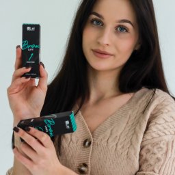 Martyna Mierzicka - MARLENS Fotografia - Kobieta prezentuje dwa czarne pudełka kosmetyków do brwi marki Bron Lift, ubrana w beżowy sweter i czarny zegarek na skórzanym pasku.