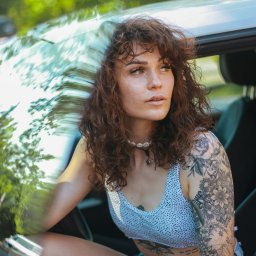 Martyna Mierzicka - MARLENS Fotografia - Portret kobiety z kręconymi brązowymi włosami i tatuażami na ramieniu, siedzącej w samochodzie, z widocznym fragmentem zieleni w tle.