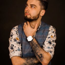 Martyna Mierzicka - MARLENS Fotografia - Mężczyzna z brodą, w kwiecistej koszuli i kraciastej kamizelce, z tatuażami na ramionach i zegarkiem na nadgarstku, spogląda w górę na ciemnym tle.