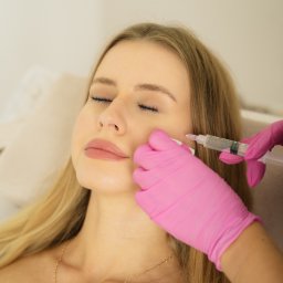 Martyna Mierzicka - MARLENS Fotografia - Blondynka z zamkniętymi oczami podczas zabiegu wypełniania zmarszczek. W dłoni różowa rękawiczka i strzykawka. Na szyi złoty łańcuszek.