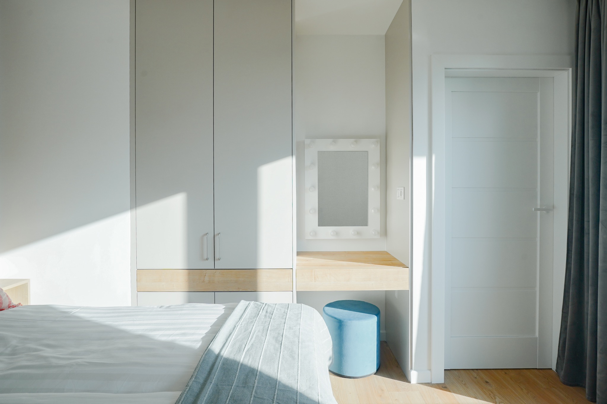 Jasna sypialnia z zabudowaną szafą, toaletką z lustrem podświetlanym żarówkami i błękitnym pufem, widoczny fragment łóżka z narzutą w paski.