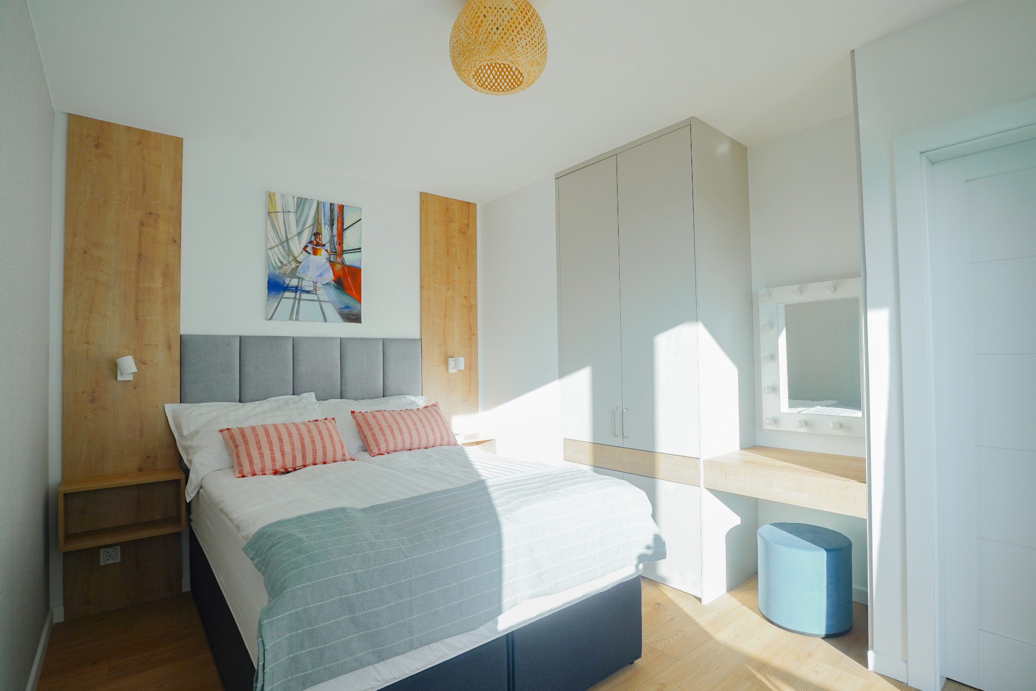 Jasna sypialnia z łóżkiem, szafą i toaletką z lustrem podświetlanym żarówkami, oświetlona promieniami słońca padającymi przez okno.