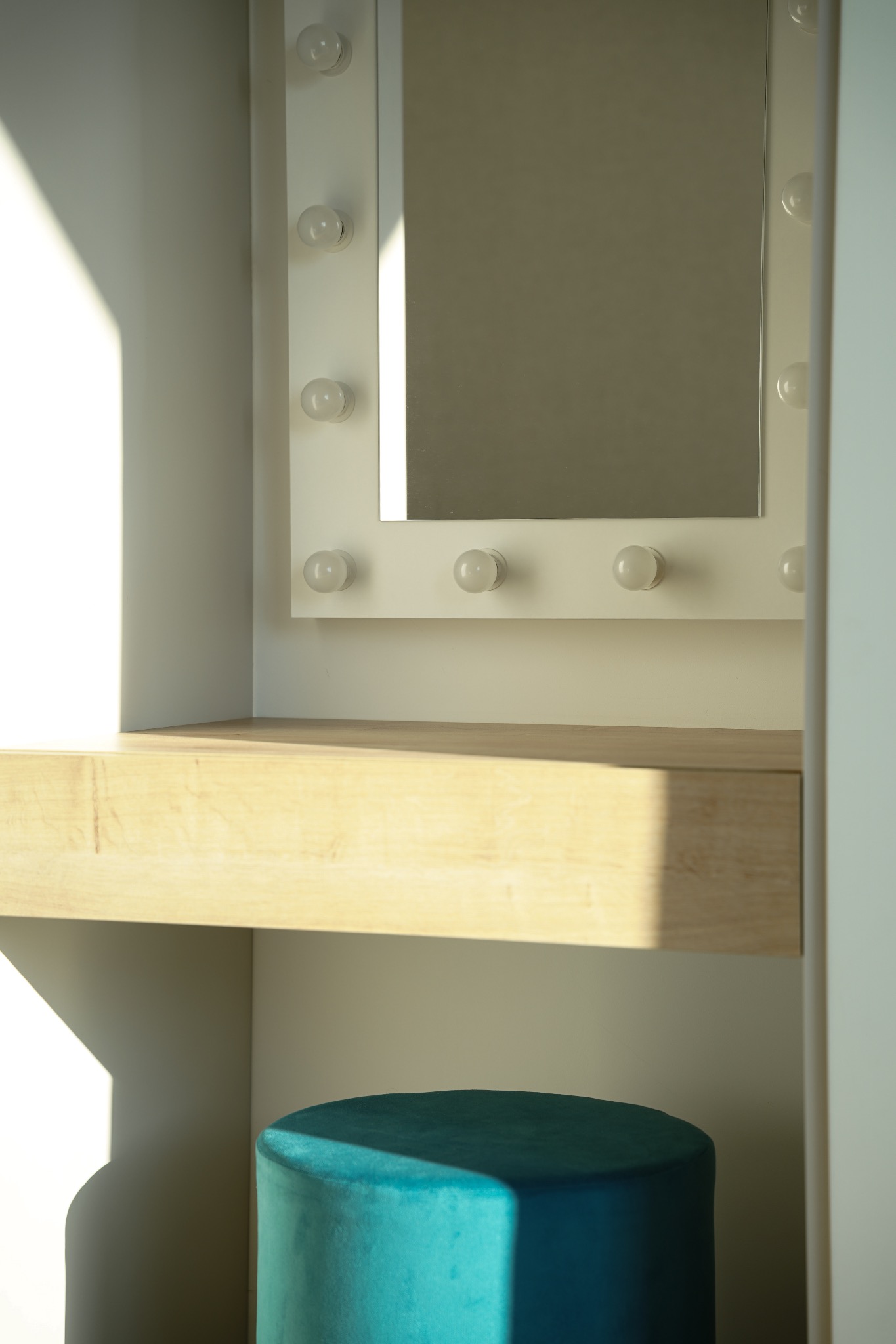 Biała toaletka z lustrem otoczonym żarówkami i turkusowym pufem, oświetlona promieniami słonecznymi.