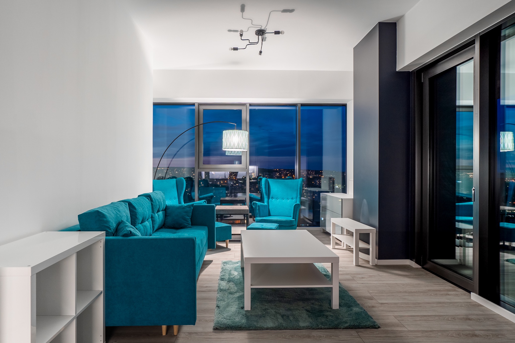 Nowoczesny salon z turkusowymi meblami wypoczynkowymi, białymi stolikami i widokiem na nocne miasto zza panoramicznych okien.