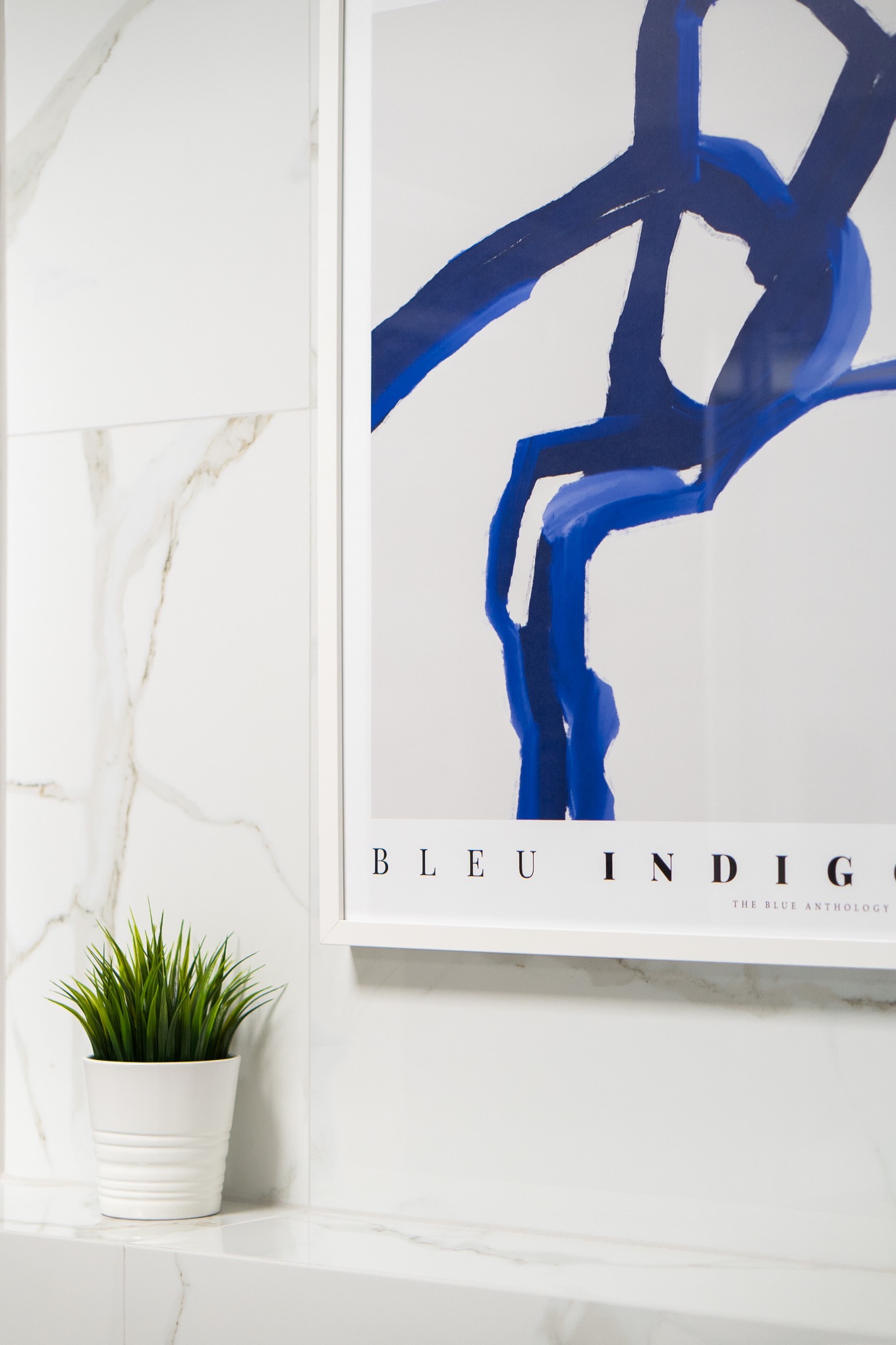 Nowoczesna łazienka z marmurowymi płytkami, na blacie biała doniczka z zieloną trawą i oprawiony plakat w kolorze indygo.