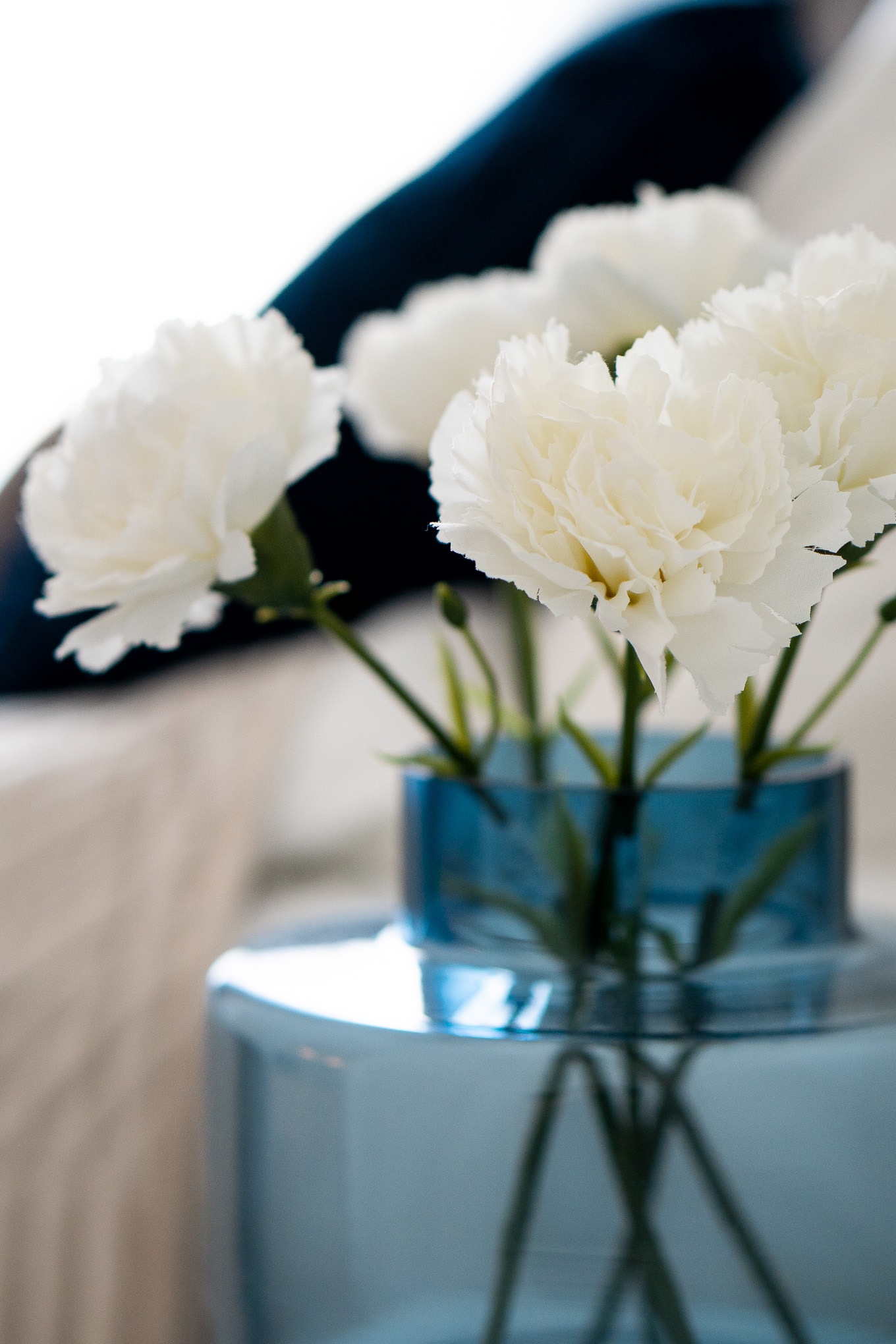 Białe goździki w niebieskim, szklanym wazonie na niebieskim stoliku, w tle beżowa sofa i granatowa poduszka.