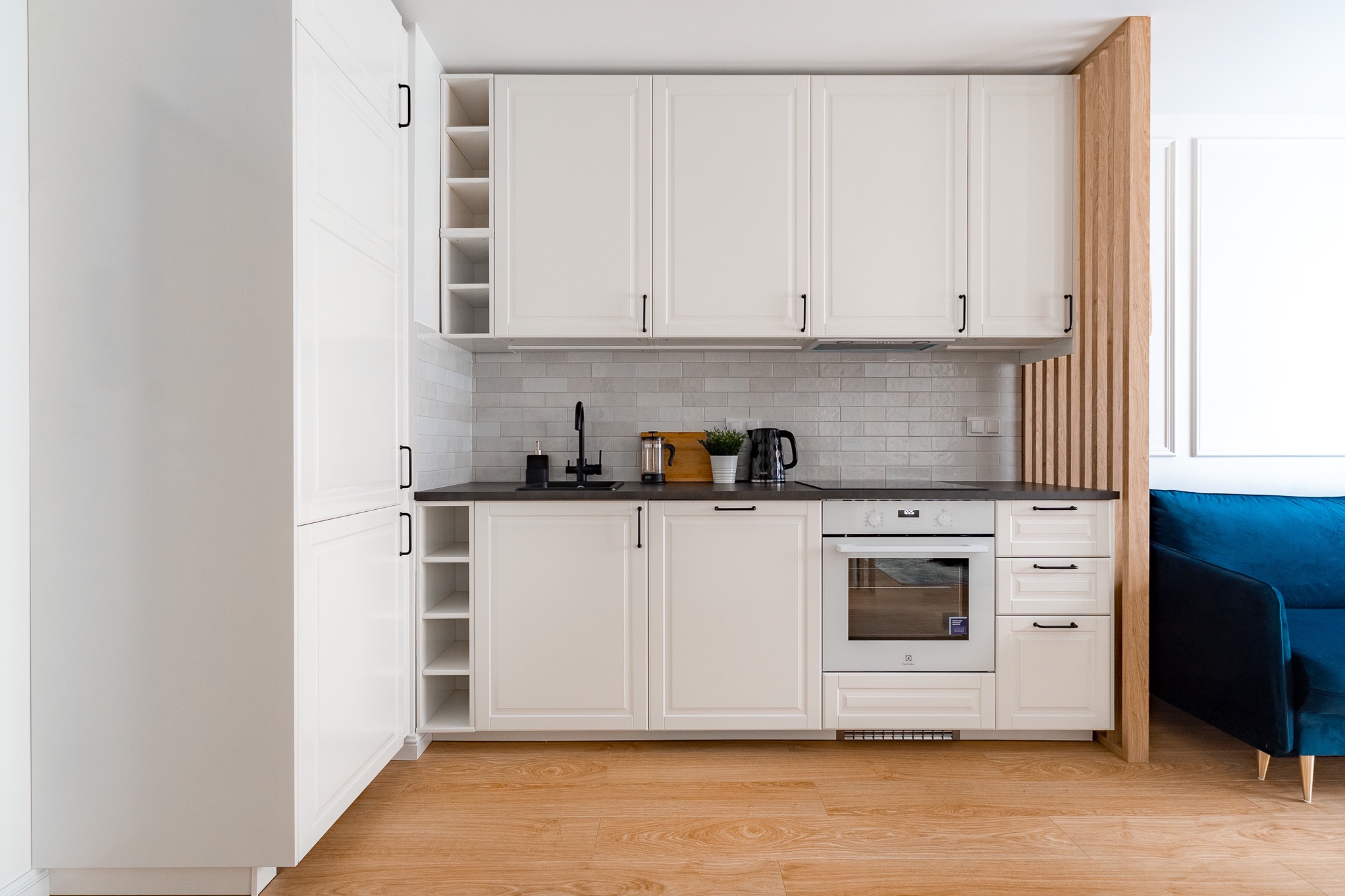 Jasna kuchnia z białymi szafkami i czarnymi uchwytami, drewniana podłoga, zlew z czarną armaturą, drewniana ażurowa ścianka działowa i fragment niebieskiej sofy.