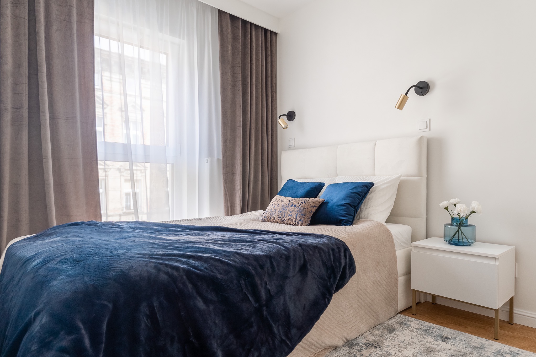 Jasna sypialnia z dużym oknem, brązowymi zasłonami i białymi firankami. Na łóżku granatowa narzuta i niebieskie poduszki. Biała szafka nocna z niebieskim wazonem i białymi kwiatami. Na ścianie...