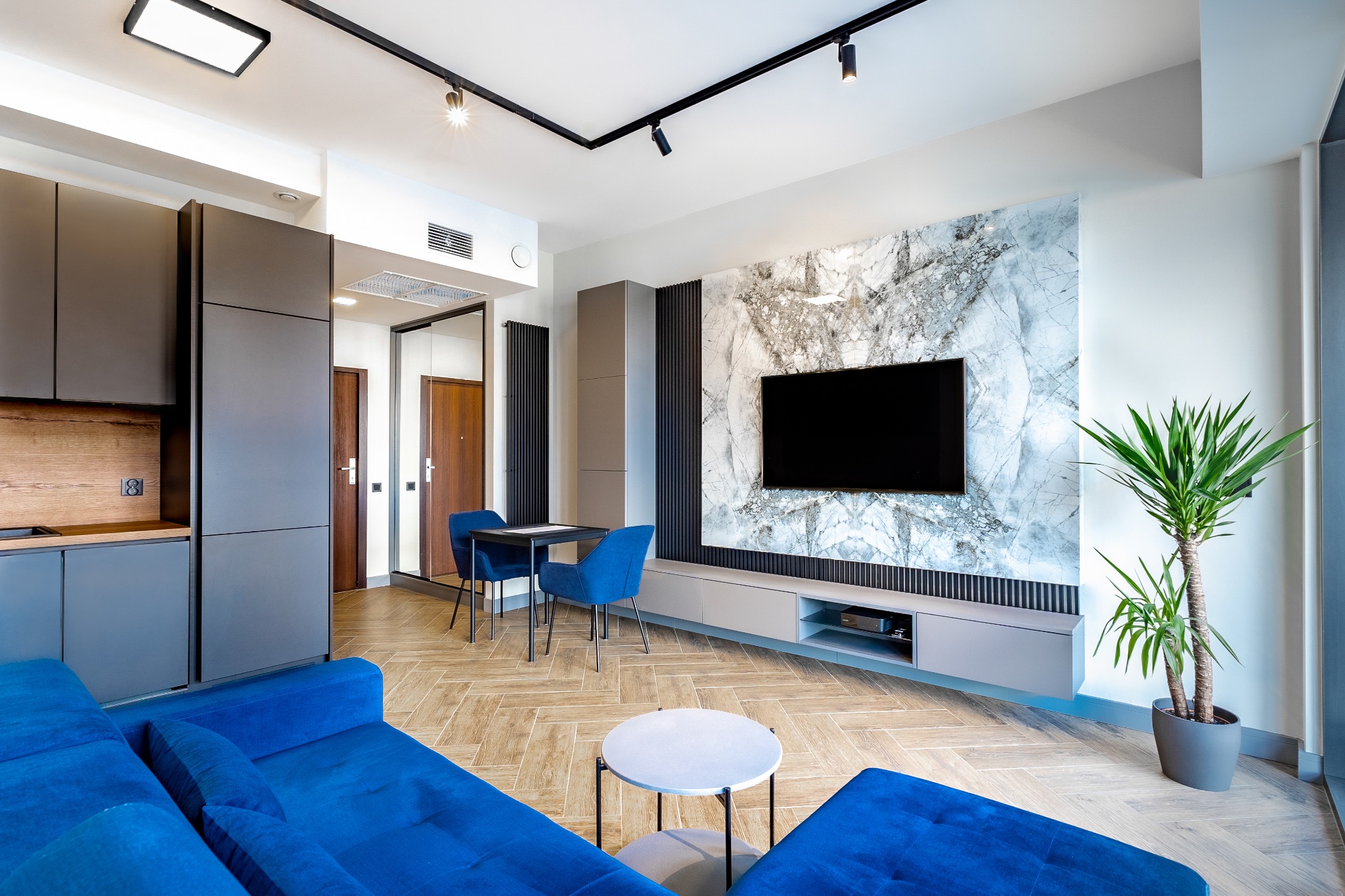 Nowoczesny salon z niebieską sofą, marmurową ścianą za telewizorem, drewnianą podłogą w jodełkę i rośliną doniczkową w szarej donicy.