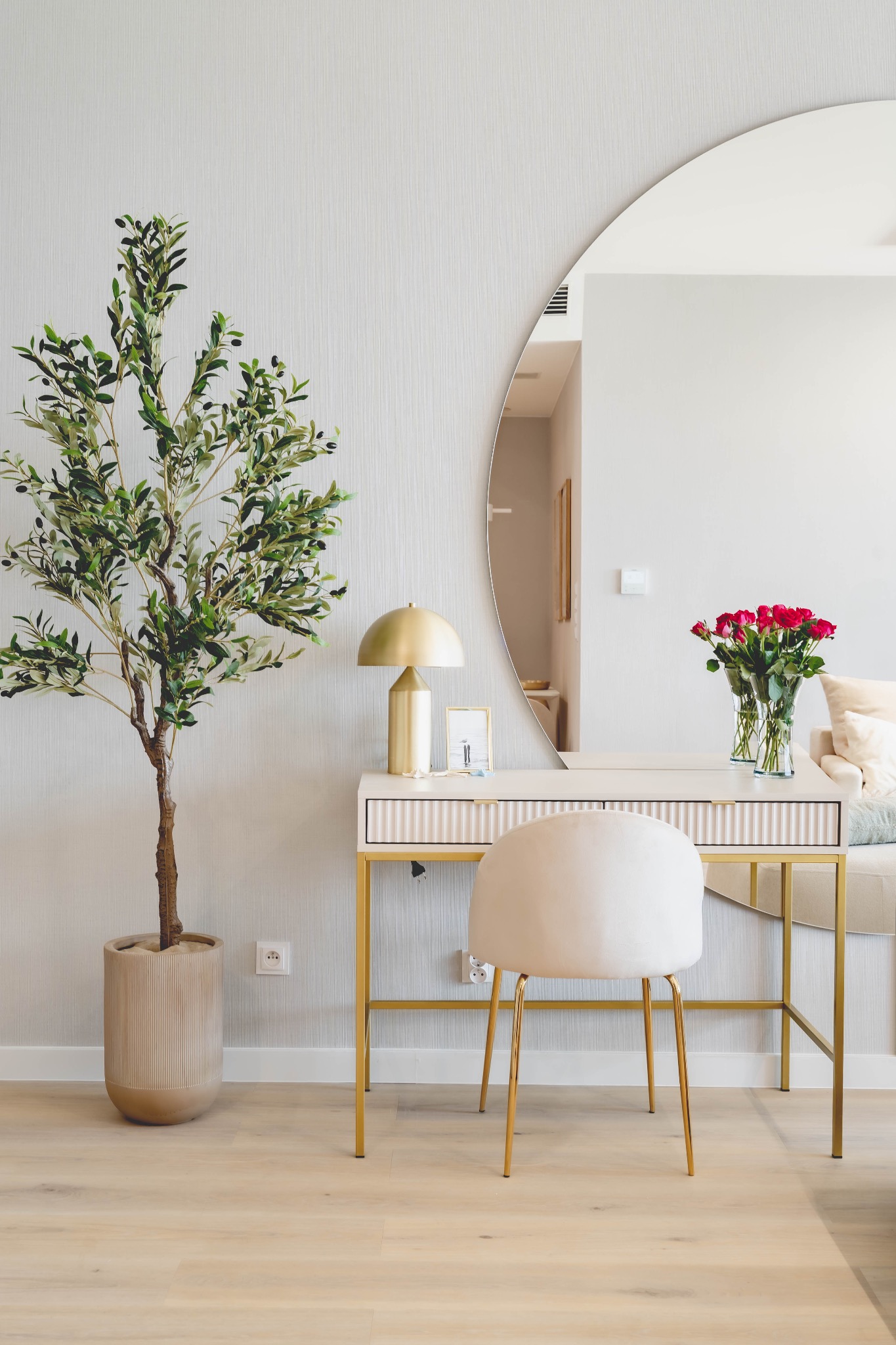 Jasne wnętrze z toaletką w stylu glamour, okrągłym lustrem i bukietem czerwonych róż. Obok drzewko oliwne w beżowej donicy i złota lampka.