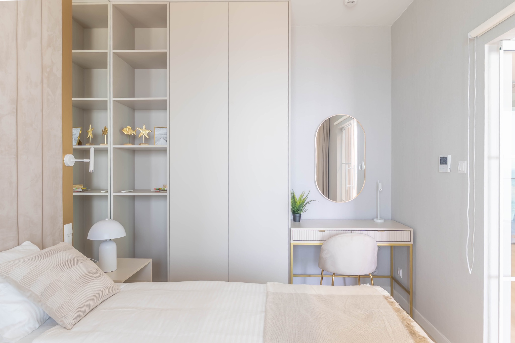 Jasne, minimalistyczne wnętrze sypialni z łóżkiem, toaletką ze złotymi nogami i dużą szafą. Neutralna kolorystyka, subtelne oświetlenie, dekoracyjne elementy.
