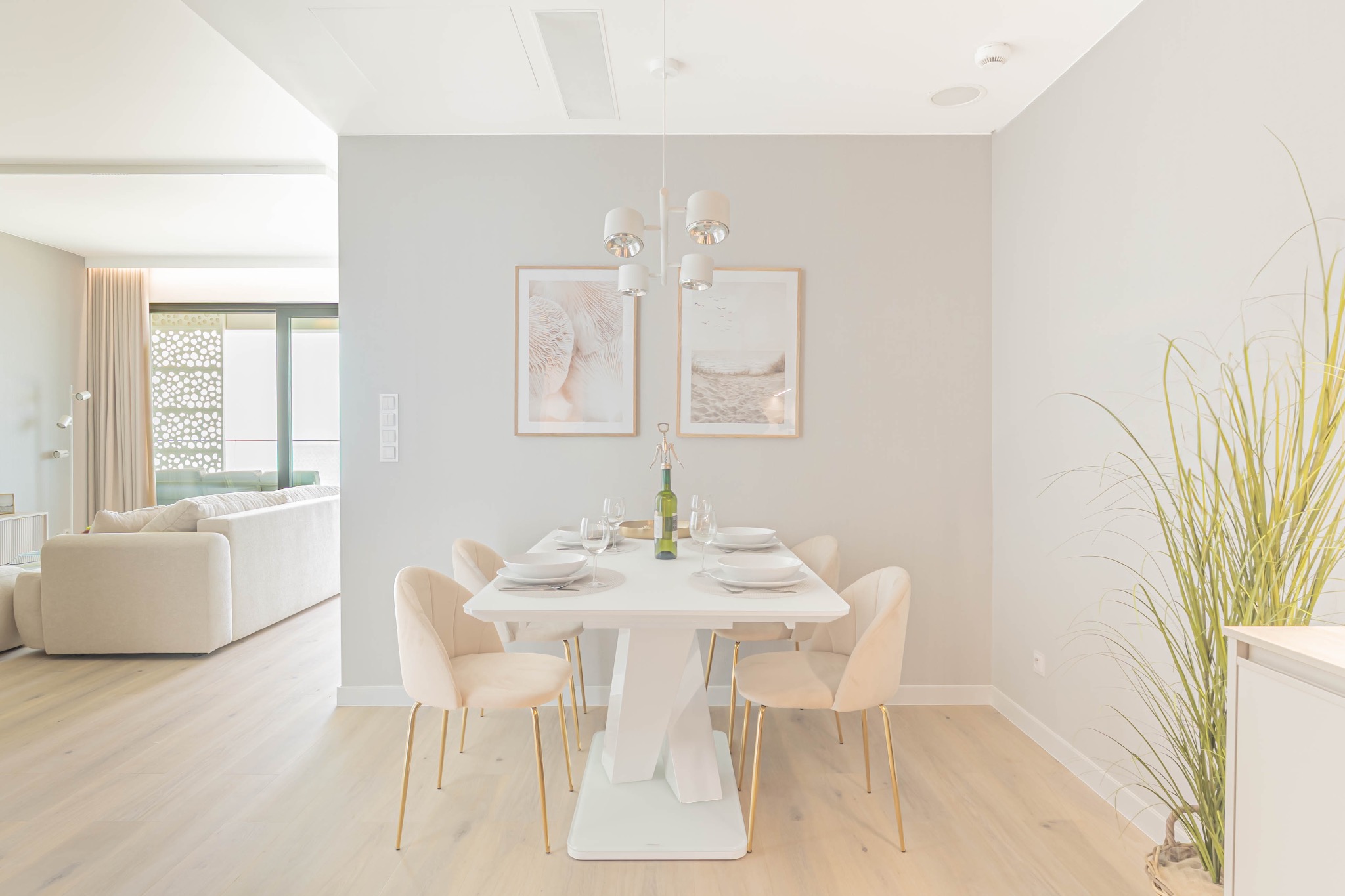 Jasne wnętrze jadalni z białym stołem, beżowymi krzesłami i dekoracjami ściennymi. W tle salon z beżową sofą i wyjściem na balkon.