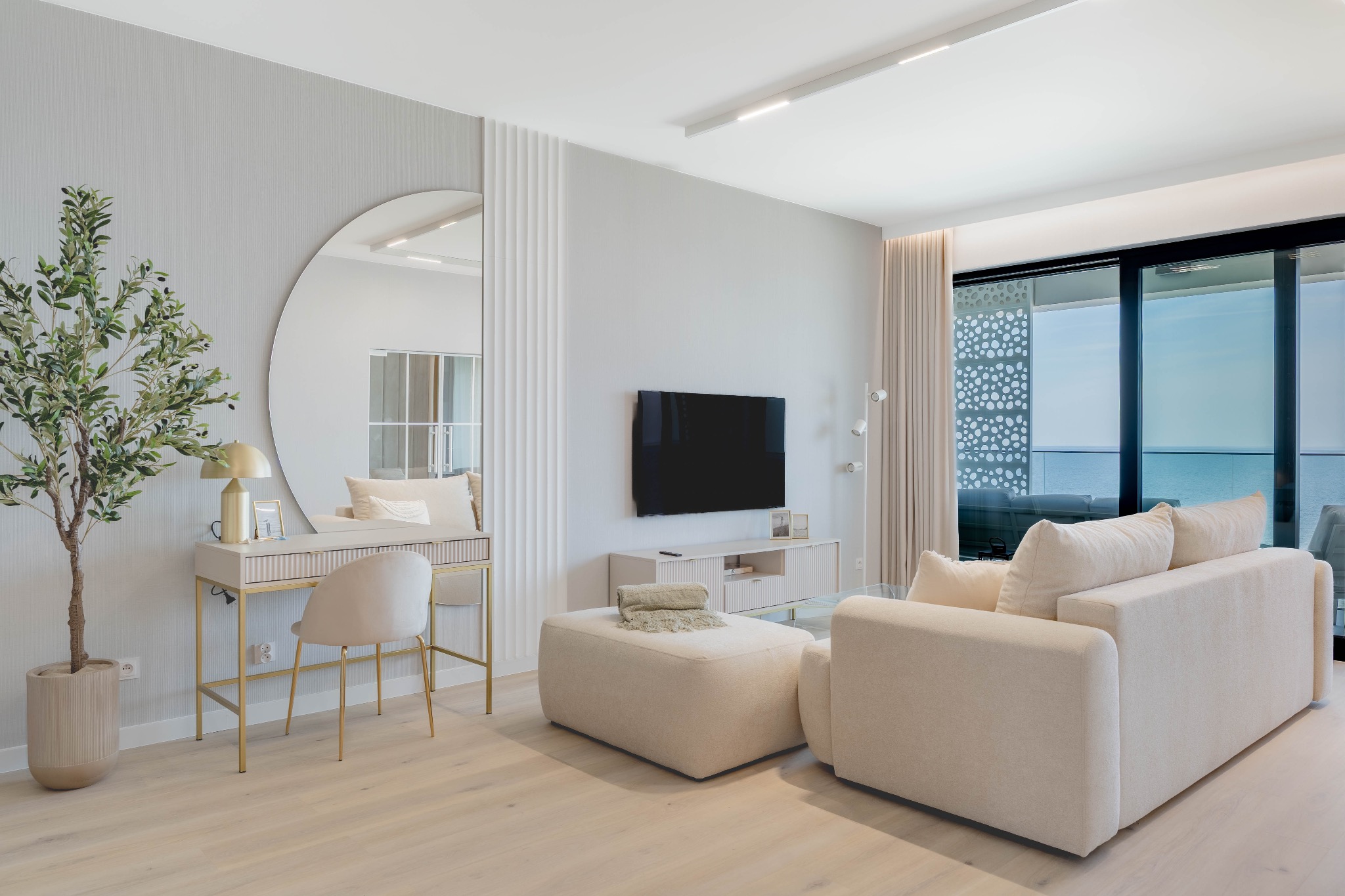 Jasne wnętrze salonu z toaletką i okrągłym lustrem, beżowa sofa, widok na morze z balkonu. Minimalistyczny design, naturalne światło.