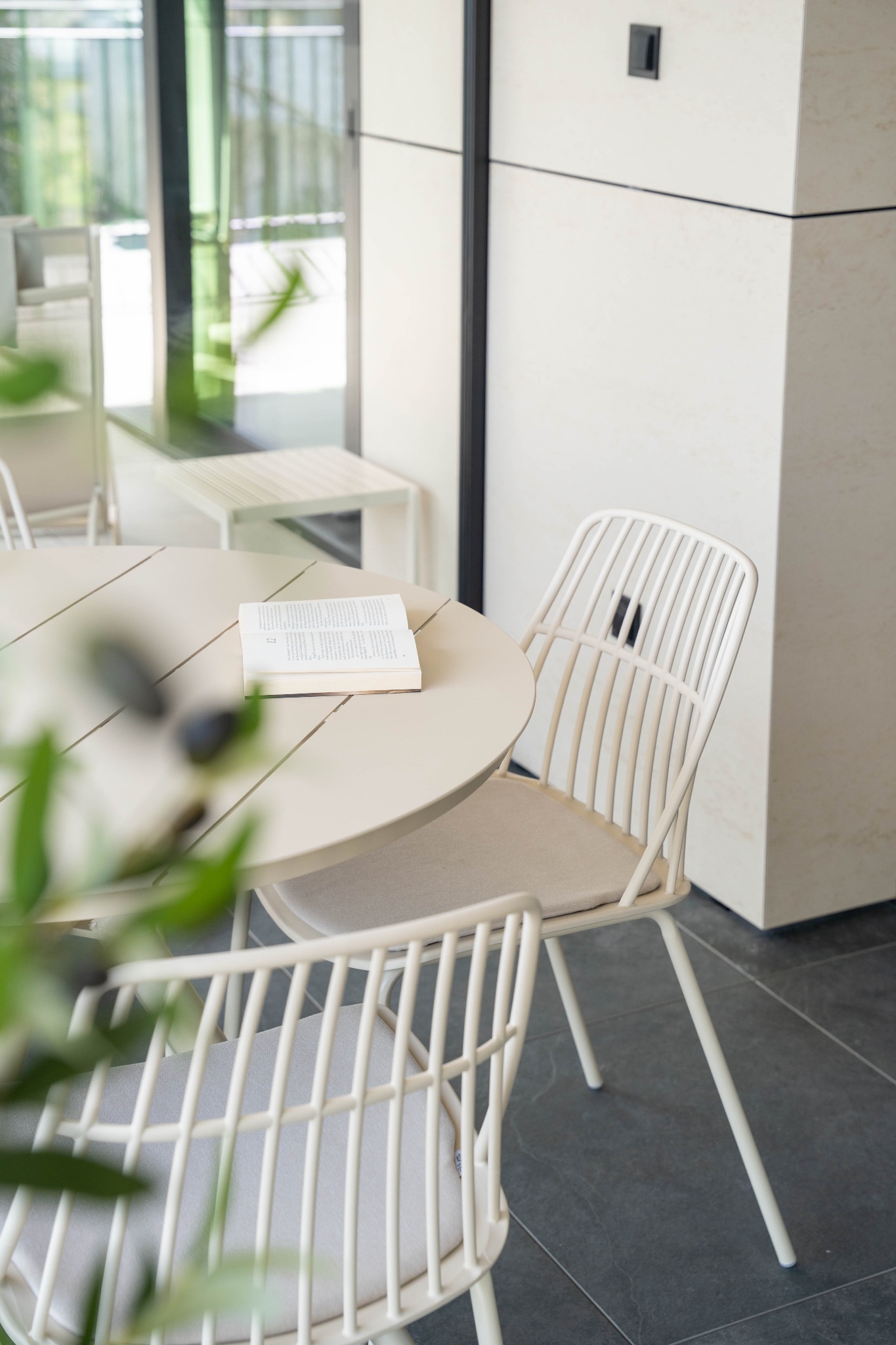 Jasne wnętrze z okrągłym stołem, otwartą książką i krzesłami o ażurowych oparciach. Minimalistyczny design, stonowana kolorystyka i naturalne światło.