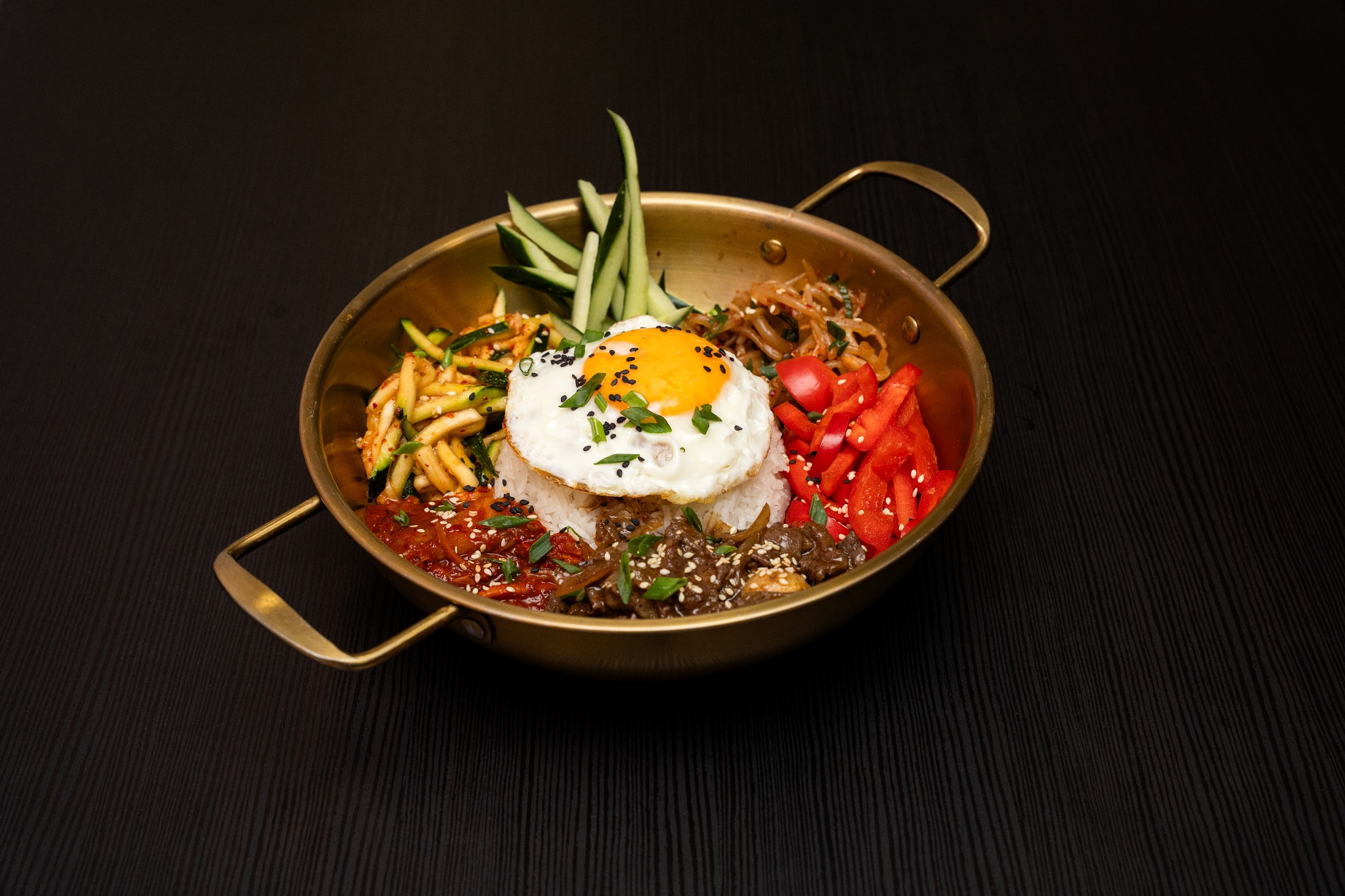Bibimbap w miedzianej misce na ciemnym tle: ryż, jajko sadzone, warzywa i mięso. Danie koreańskie, z bliska, ostre światło, widoczne detale.