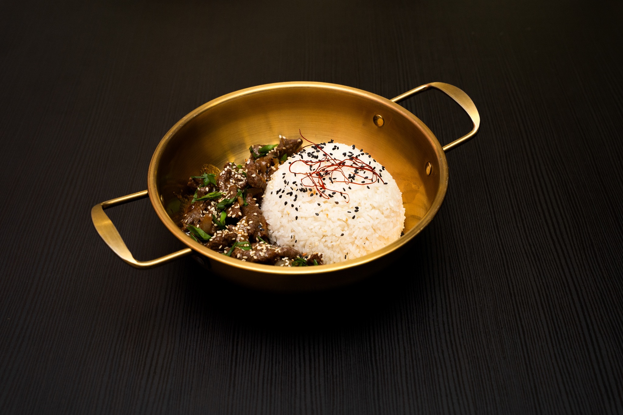 Danie azjatyckie: wołowina teriyaki z ryżem w złotej misce na ciemnym tle. Ryż posypany sezamem, całość oświetlona punktowo.