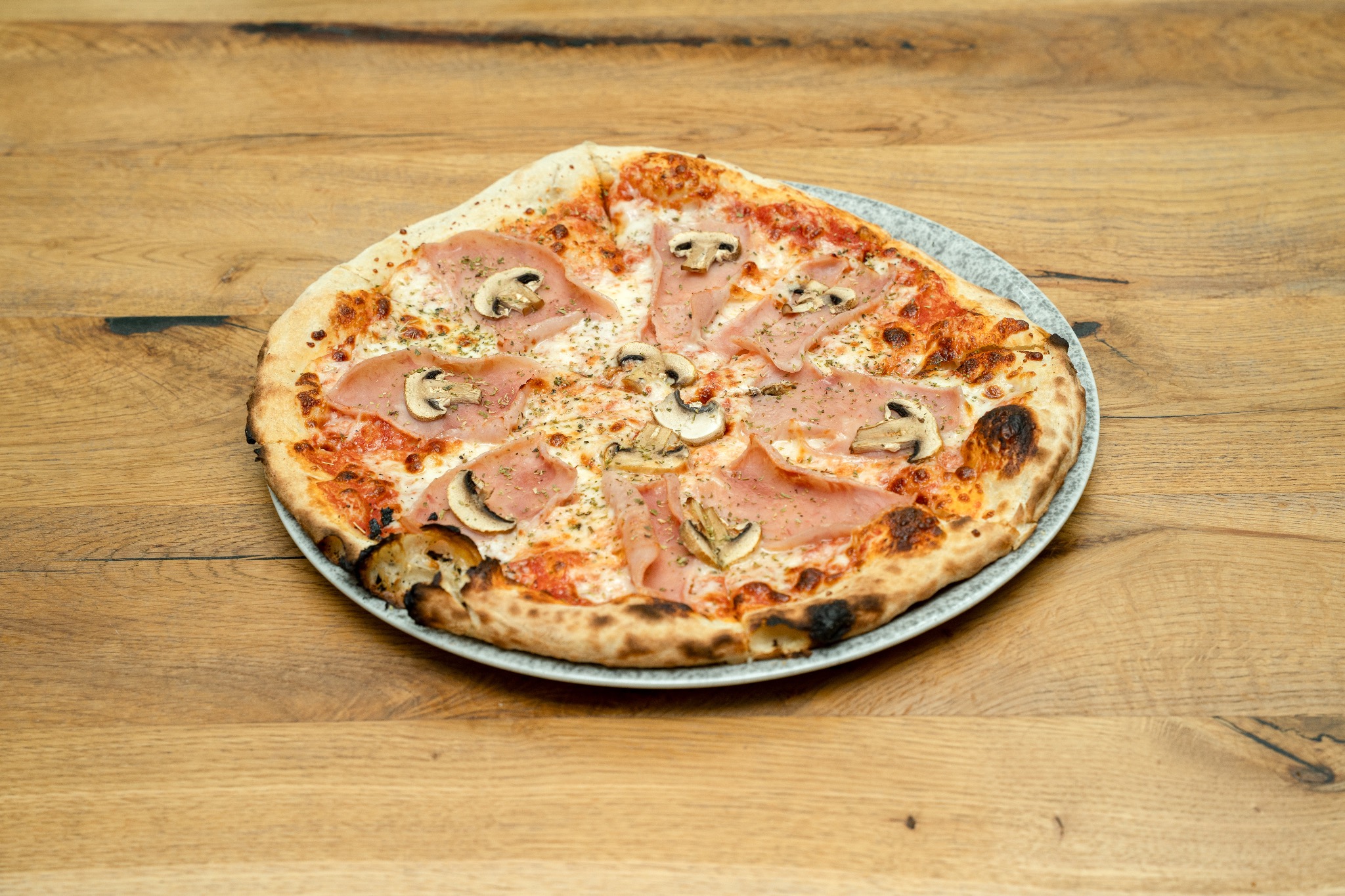 Pizza z szynką i pieczarkami na talerzu z kamionki, na drewnianym blacie. Widoczne przyprawy i przypieczone brzegi ciasta.