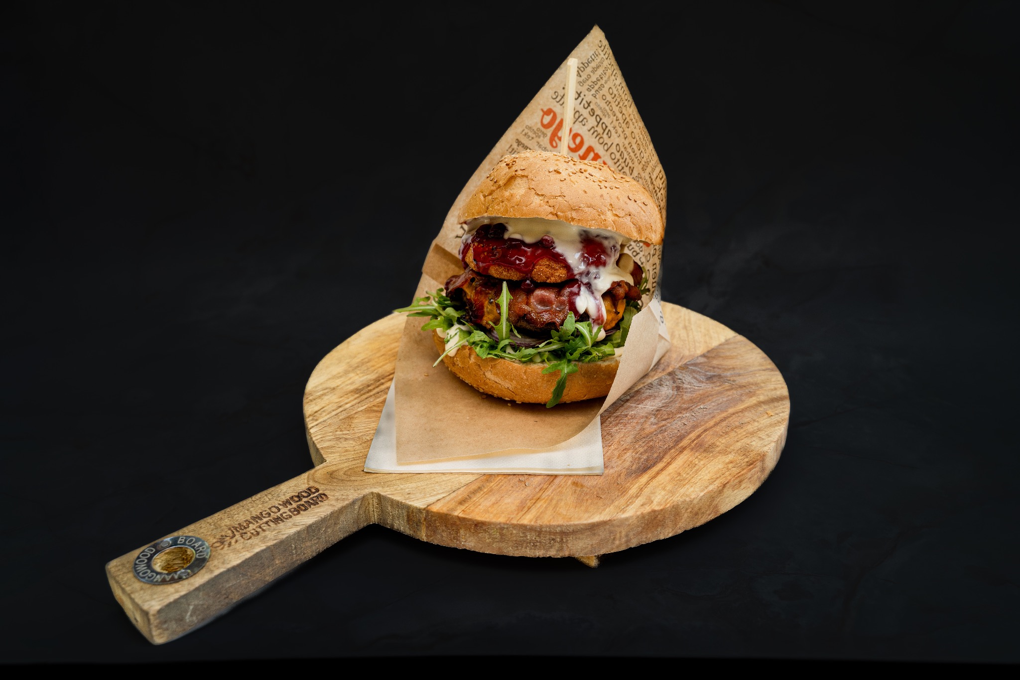 Apetyczny, dwupoziomowy burger z sosem, świeżymi ziołami i sezamową bułką, podany na drewnianej desce do krojenia z uchwytem na czarnym tle.
