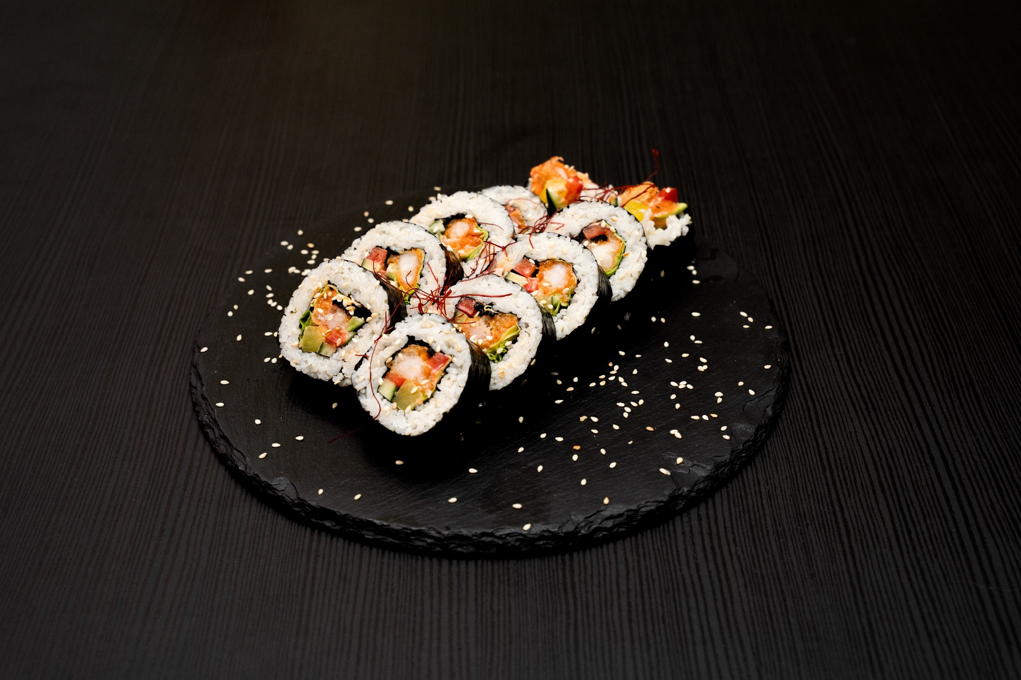 Sushi z krewetkami na czarnym talerzu z łupka, posypane sezamem, na tle ciemnego, drewnianego stołu. Ujęcie z góry, ostre światło.