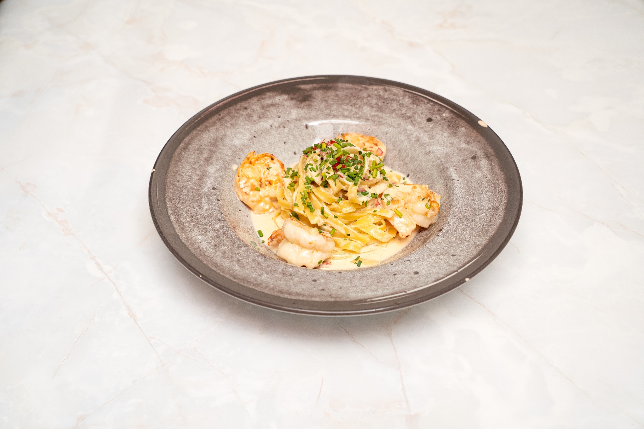 Aromatyczne tagliatelle z krewetkami i sosem śmietanowym, posypane świeżym szczypiorkiem, serwowane na talerzu z szarej ceramiki na marmurowym blacie.