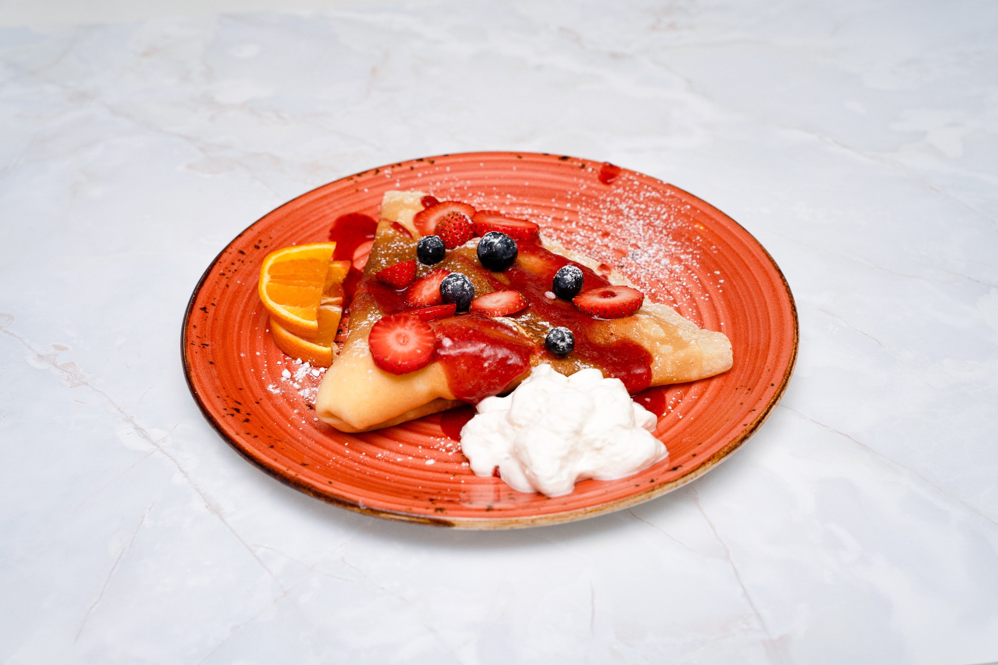 Apetyczny naleśnik na pomarańczowym talerzu, udekorowany truskawkami, borówkami, sosem i bitą śmietaną. Całość posypana cukrem pudrem, z boku plaster pomarańczy.