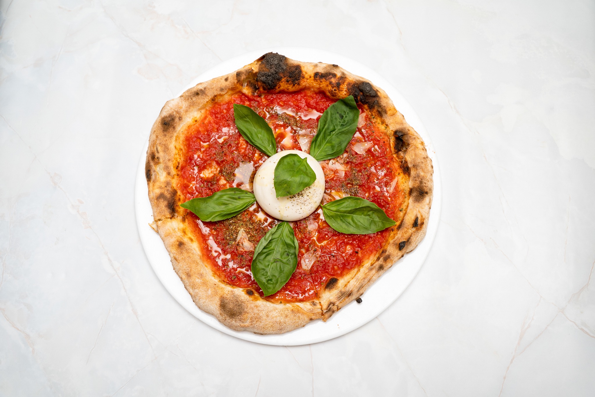 Pizza Margherita z bazylią i burratą na marmurowym tle. Ciasto z lekko przypalonym brzegiem. Widok z góry, ostre światło podkreśla świeżość składników.