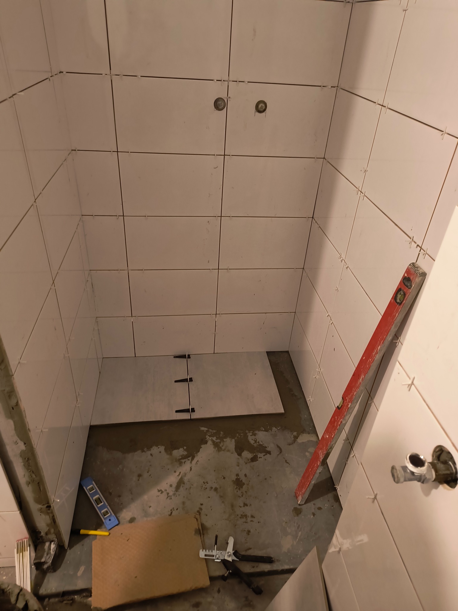 Wnętrze kabiny prysznicowej w trakcie układania białych płytek, widoczne krzyżyki dystansowe, poziomica, narzędzia glazurnicze i fragment instalacji wodnej.