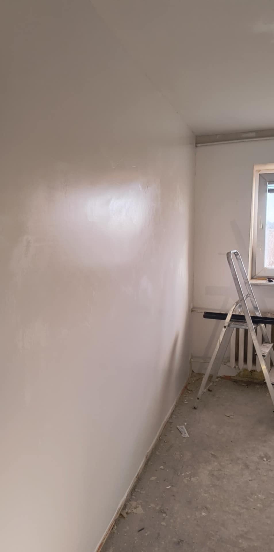 Świeżo nałożona gładź na ścianie w rogu pokoju, widoczne ślady szpachlowania, drabina aluminiowa oparta o ścianę przy oknie, surowy beton na podłodze.