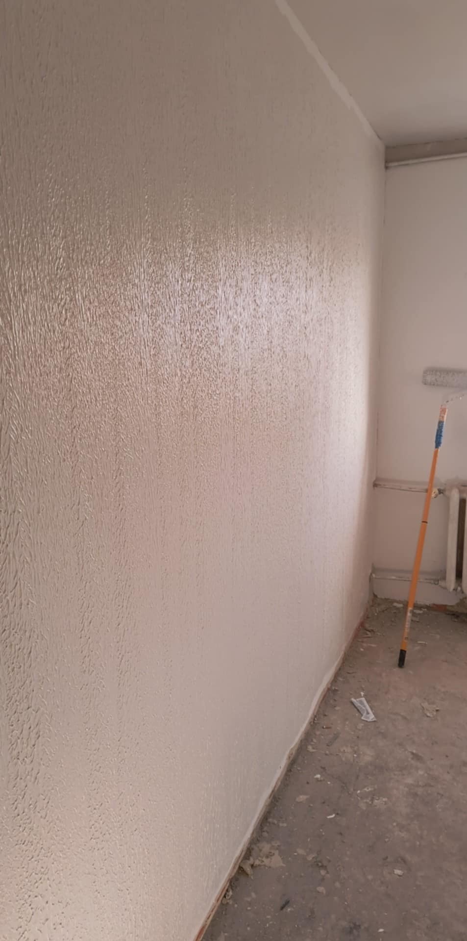 Świeżo pomalowana ściana z fakturą, widoczny wałek malarski oparty o ścianę, surowy betonowy podkład w pomieszczeniu.