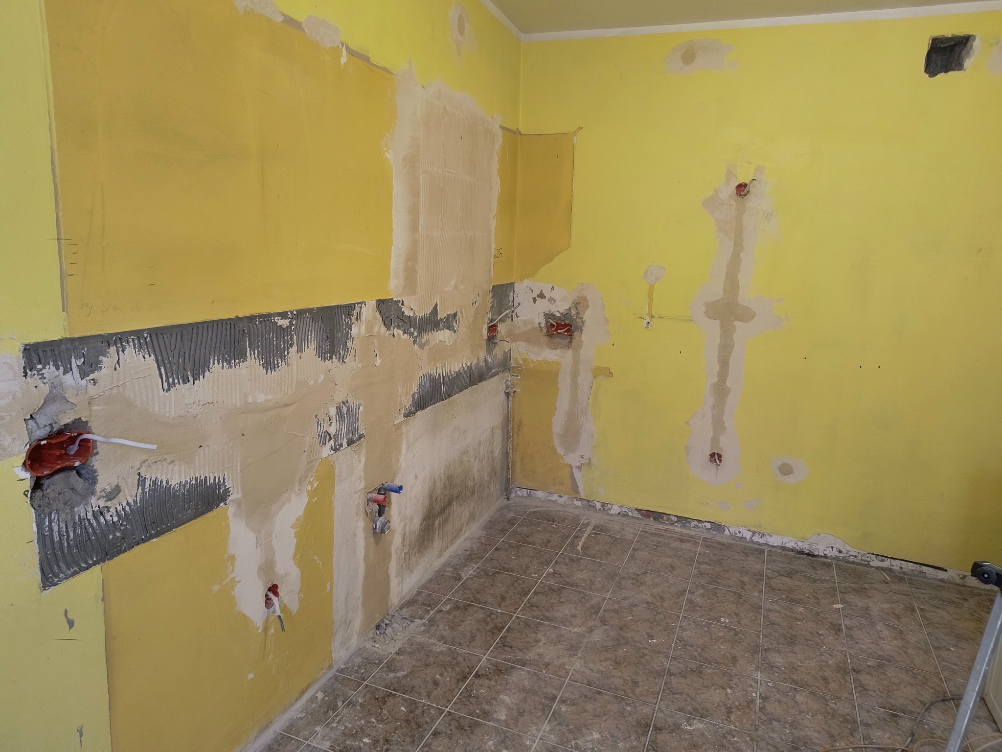 Żółta ściana w trakcie przygotowania do położenia płytek, widoczne fragmenty z nałożonym klejem i instalacjami hydraulicznymi, podłoga wyłożona brązowymi płytkami, ściany częściowo zagruntowane.