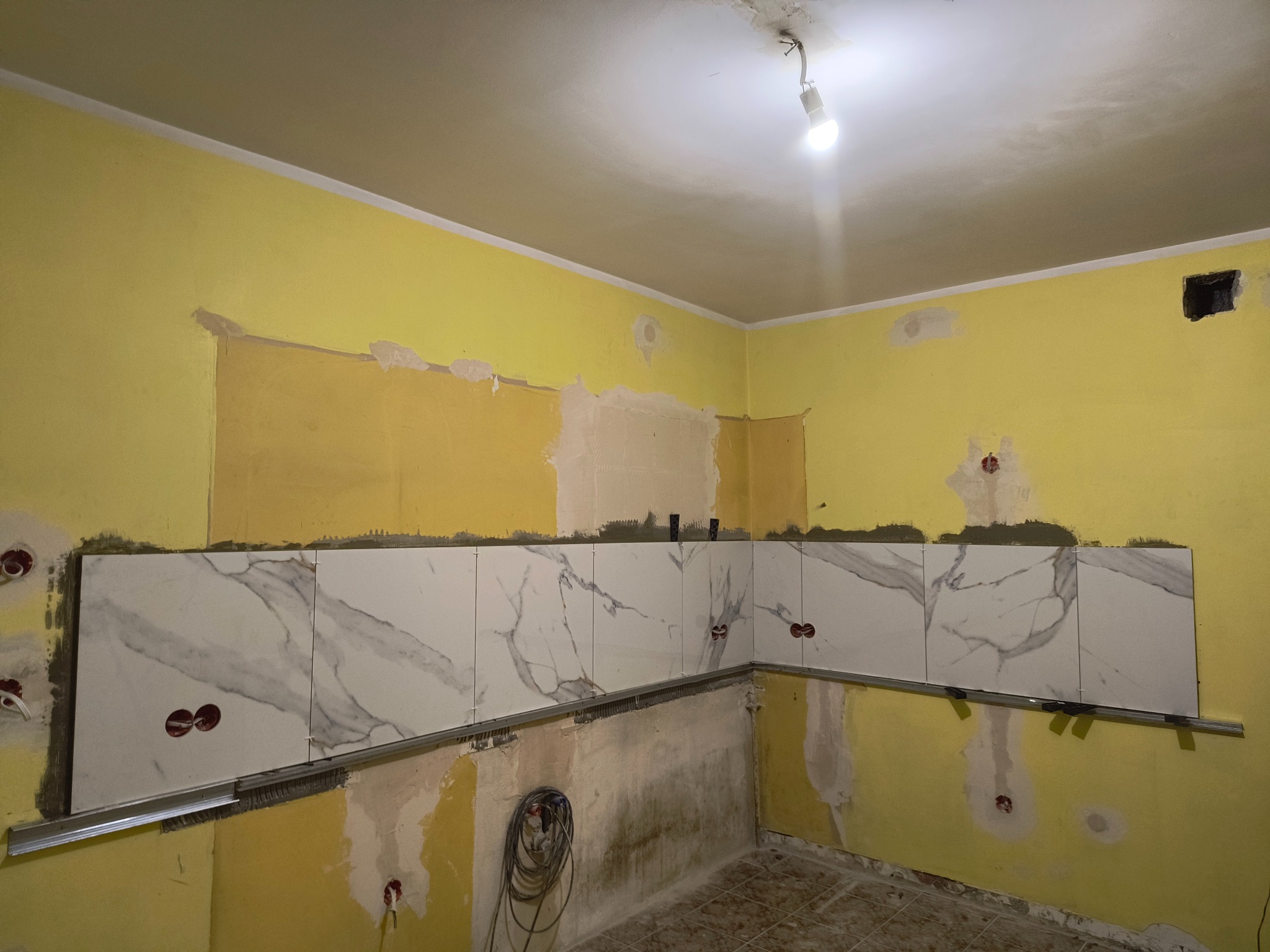 Żółta ściana w trakcie remontu kuchni z częściowo położonymi białymi płytkami imitującymi marmur, zamocowanymi na szynie, widoczne gniazdka elektryczne i wiszący kabel.